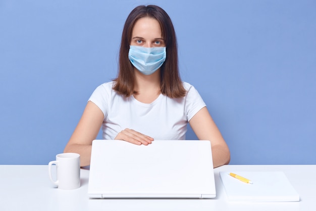 Tiro al coperto di giovane donna che lavora da casa con il computer portatile, indossando la maschera protettiva, copywriter essendo pronto a lavorare, ragioniere ha lavoro a distanza, seduto al tavolo bianco. libero professionista durante la quarantena.