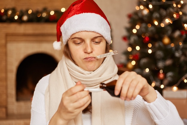 Indoor sot of ill woman wearing santa claus hat, being wrapped in warm scarf, sitting near Christmas tree, pouring syrup, needs treatment, having flu symptom, celebrating new year alone.