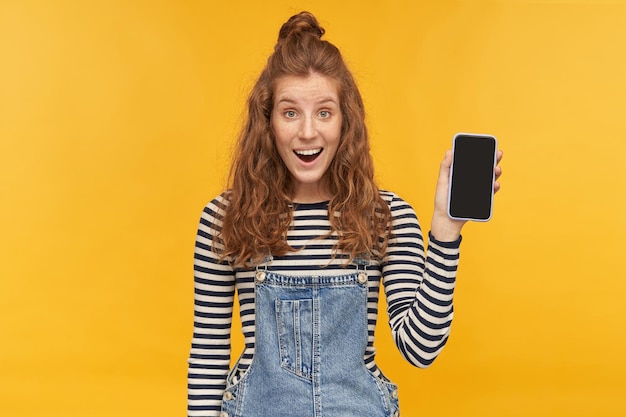 Indoor studio shot of young ginger female with long red curly hair smiles broadly with positive facial expression, shows black blank phone display into camera. isolated over yellow background