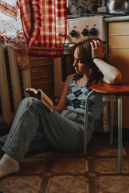 Indoors a teenage girl sits on the floor in the kitchen and reads a book