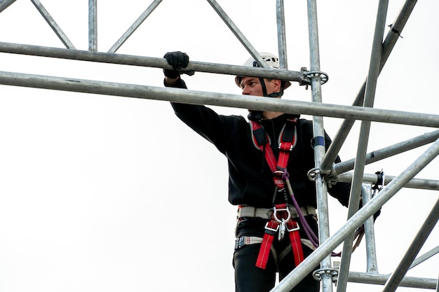 An industrial climber wearing a helmet and a protective belt is tied with a rope to a support at a high altitude A specialist assembles a stage structure from a modular system of metal scaffolding
