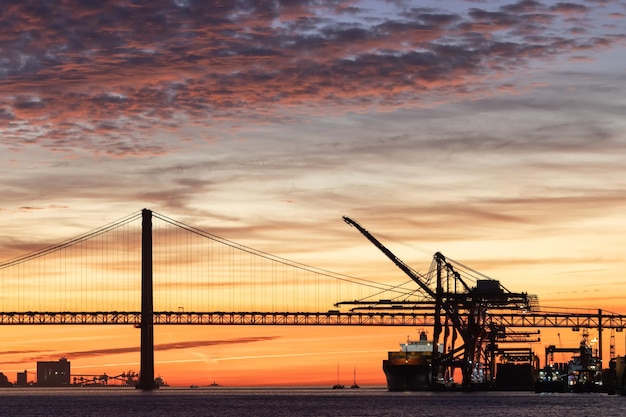 Industrial cranes a suspension bridge and a tourist liner sailing along the river at sunset