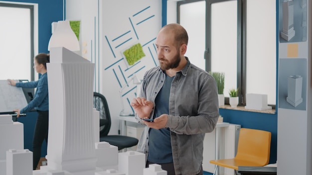 Industrial engineer designing construction plan with building model and smartphone. Architect using mobile phone and maquette on table to work on project development, to build property.