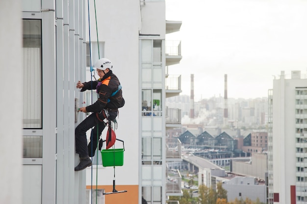 Промышленный альпинист висит над жилым домом во время мытья фасадного остекления. Рабочий висит на стене дома. Концепция городских работ. Копировать пространство