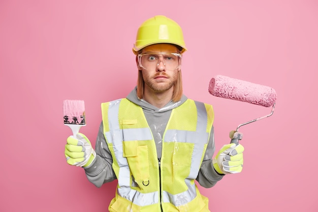 Industrial work repair concept. Serious male builder on construction site holds building tools wears protective safety clothes ready to paint walls isolated on pink wall. Professional repairman