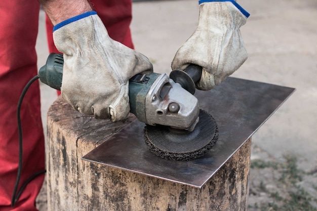 Un operaio industriale in tuta rossa con le mani in guanti da lavoro macina un pezzo di lamiera d'acciaio con una vecchia smerigliatrice