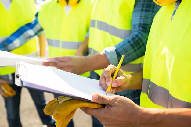 industrie, gebouw, papierwerk en mensenconcept - close-up van bouwers in goed zichtbare vesten die naar klembord schrijven