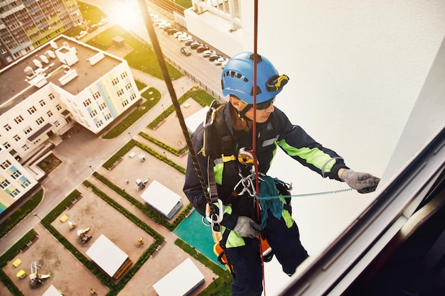 Industry urban works rope access laborer on wall of house top\
view of industrial mountaineering worker in uniform hangs over\
residential building at background