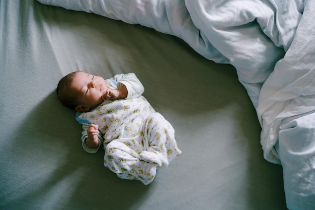 Photo infant in the crawlers sleeps on the bed arms and legs bent top view
