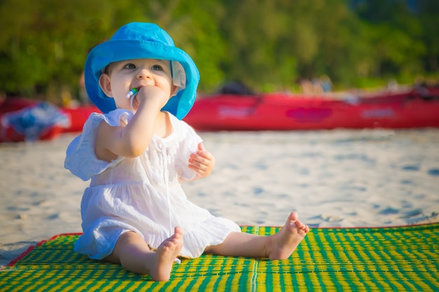 Младенческая девочка тянет в рот веревку из платья, сидя на песчаном пляже в первый раз и впечатлена