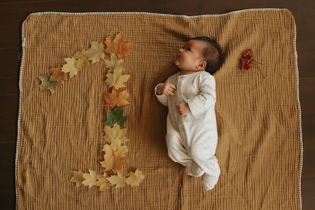 An infant is lying on a muslin blanket near maple leaves lying in the shape of 1 and staring to the left. A cute baby girl in one-piece clothing is celebrating her 1 monthly birthday.