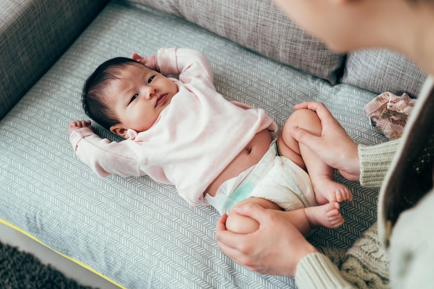 infant lying on sofa is feeling uncomfortable with wet diaper. pov shot baby raising both hands and frowning at camera while her mom is holding her feet.