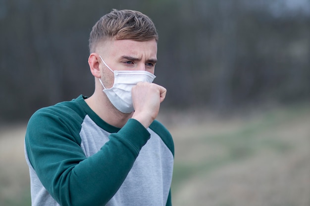 Infected guy, young sick ill man coughs, holds his hand on chest, feeling bad in protective sterile medical mask on face outdoors. Air pollution, virus, pandemic coronavirus concept. Covid-19