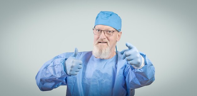 Infectious disease doctor with a gray beard in a blue protective overalls and rubber gloves shows a thumbs up