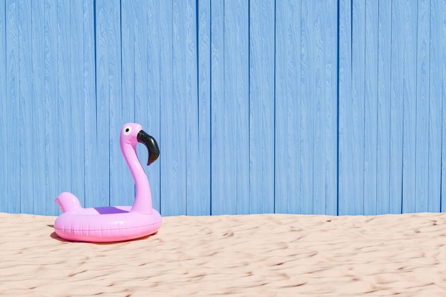 Надувной розовый фламинго на песчаном пляже с синим деревянным фоном