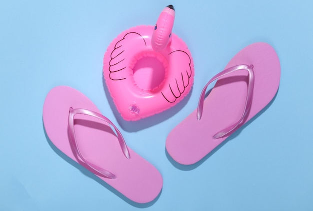 Inflatable pink flamingo with flip flops on blue sunny background. summer, beach vacation concept. minimalism. top view.