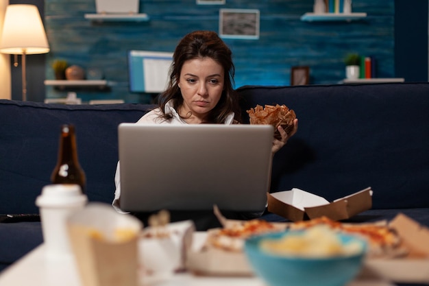 Influencer checking social media on laptop eating a tasty\
takeout burger and fast food delivery menu in living room. content\
creator watching online series while having takeaway\
hamburger.