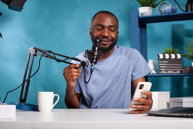 Influencer having questions and answers session using microphone in recording desk in vlogging studio. African american vlogger reading live text messages from fans looking at smartphone screen.