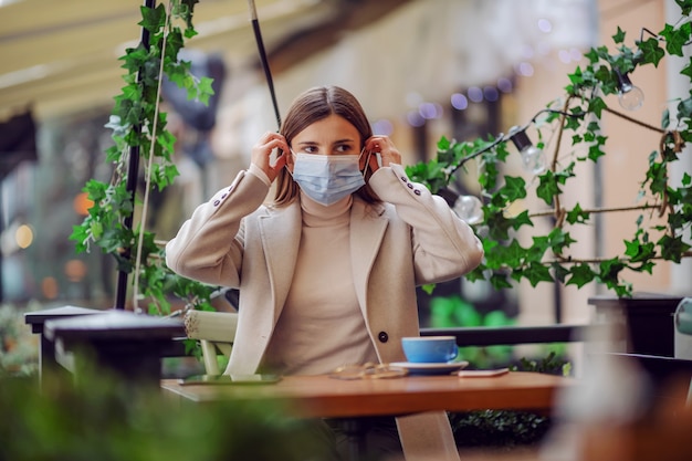 Influencer sitting in cafe and putting face mask during corona outbreak. It is forbidden to stay in public without mask.