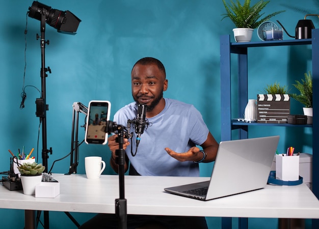 Influencer sitting down at desk with laptop in vlogging studio\
smiling in front of recording smartphone holding microphone.\
content creator interacting with fans looking at live video podcast\
setup.