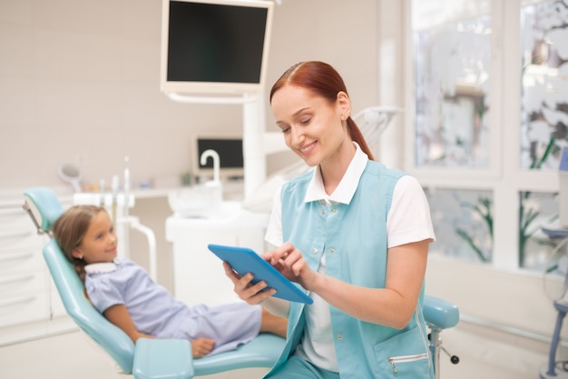Information on tablet. Red-haired child dentist smiling while filling information on tablet