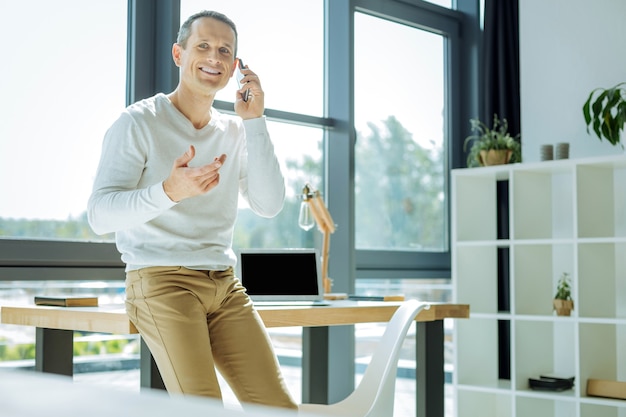 Informele sfeer. Aantrekkelijke vrolijke aardige man leunend op de tafel en praten aan de telefoon terwijl hij zich op zijn gemak voelt in zijn kantoor