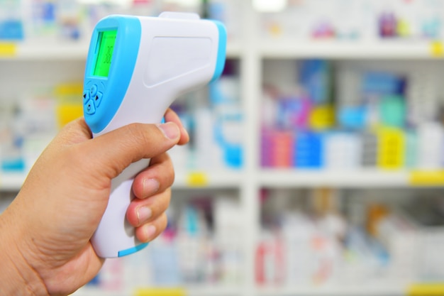 Infrared medical thermometer gun in hand on medicine shelf background.