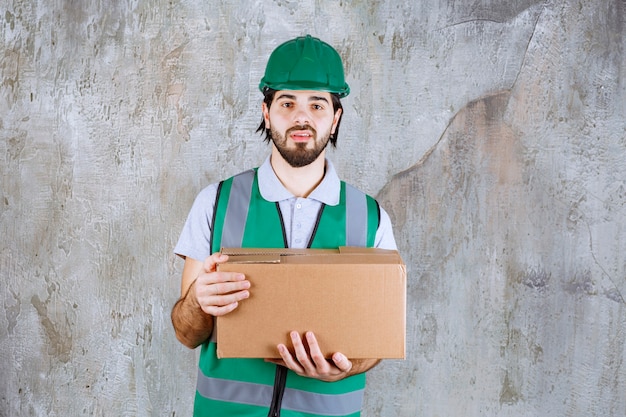 Ingenieur in gele uitrusting en helm met een kartonnen doos en ziet er verward en attent uit.