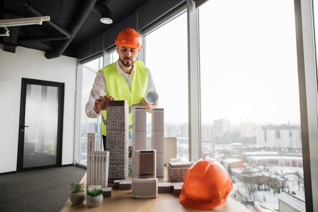 Ingenieur man in helm en reflecterend vest staande in de buurt van tafel met architectuurontwerp van de stad