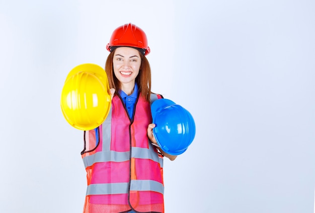 Ingenieursmeisje in uniform dat blauwe en gele helmen vasthoudt en een keuze maakt.