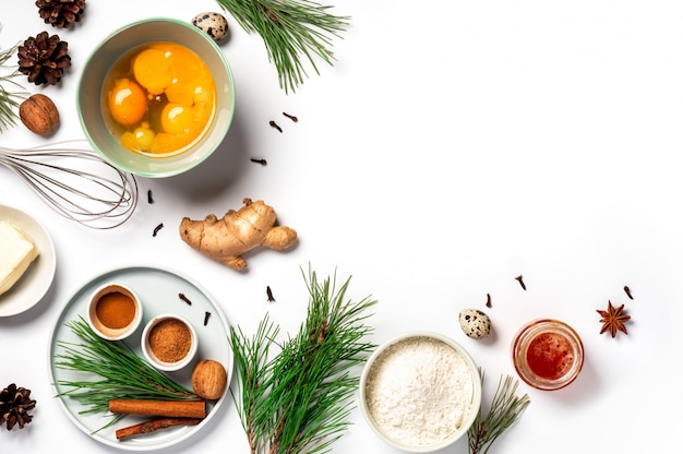 Ingrediënten voor het bakken van de gemberkoekjes van Kerstmis op een witte achtergrond, exemplaarruimte. Bloem, eieren, gember, kruiden, boter, groene pijnboomtakken en kegels op de tafel, plat liggend.