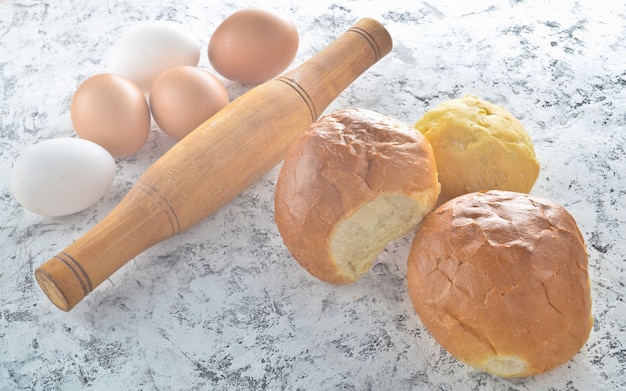 Ingrediënten voor het koken van broodjes thuis. Eieren, deegroller, broodjes op een witte betonnen tafel