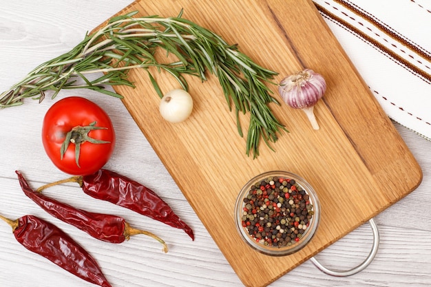 Ingrediënten voor het koken van vlees of vis. Knoflook, piment pepers in glazen kom, chili peper, tomaat, ui en rozemarijn op een houten snijplank. Bovenaanzicht.