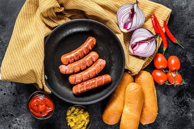 Ingrediënten voor het maken van zelfgemaakte hotdogs. Worstjes in de pan, vers gebakken broodjes, mosterd, ketchup, komkommers