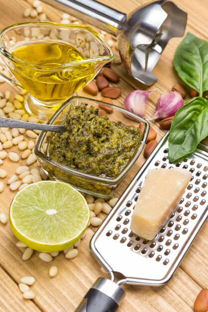 Ingrediënten voor huisgemaakte pesto. Parmezaanse kaas op rasp. pestosaus in glazen beker. Citroen en bladeren, basilicum en olie. Knoflook en pijnboompitten. Metalen shredder. Bovenaanzicht. Lichte houten achtergrond.