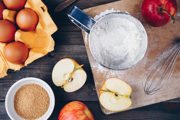 Ingredients for apple pie cooking with fresh red apples flour eggs brown sugar and spices on a rustic wooden background