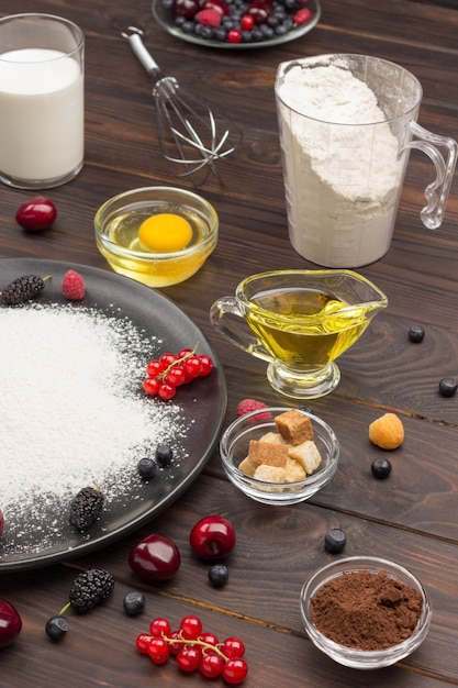 Ingredients for baking berry pie. flour in black plate, cocoa\
powder. measuring cup with flour, glass of milk, broken egg and\
salt, metal whisk on table. dark wooden surface. top view