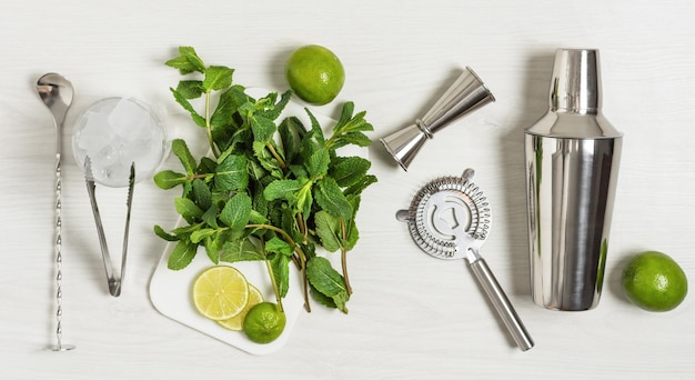 Ingredients and bar utensils for making mojito cocktail with ice cubes fresh mint and lime on white