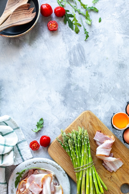 Ingredients for cooking on a gray concrete background. A bunch of fresh green asparagus, eggs, bacon, mushrooms, tomatoes, arugula. Top view. Copy space