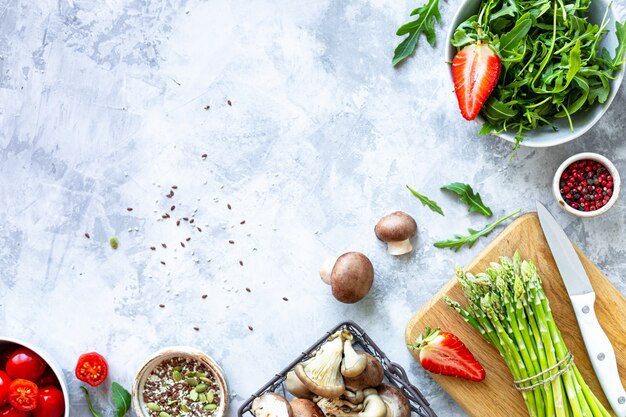 Ingredients for cooking on a gray concrete background. A bunch of fresh green asparagus, strawberries, mushrooms, tomatoes, arugula. Top view. Copy space