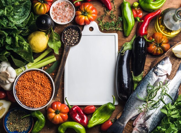 Ingredients for cooking healthy dinner Raw uncooked seabass fish with vegetables grains herbs and spices over rustic wooden background white ceramic board in center