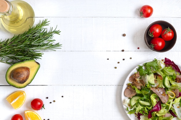 Ingredients for cooking with copy space. Bowl of fresh salad, rosemary, lemon, spices and oil on white wooden table. Food background. Healthy eating concept.