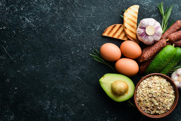 Ingredients of healthy nutrition Breakfast preparation Eggs oatmeal sausages and fresh vegetables on a black stone background