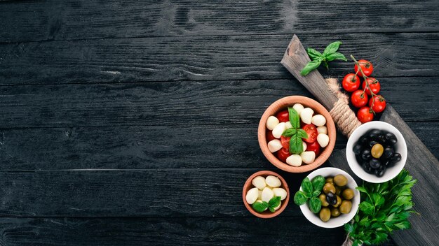 Ingredients for Italian caprese salad Mozzarella cheese cherry tomatoes basil leaves olives oil pepper On a black wooden background Free space for text