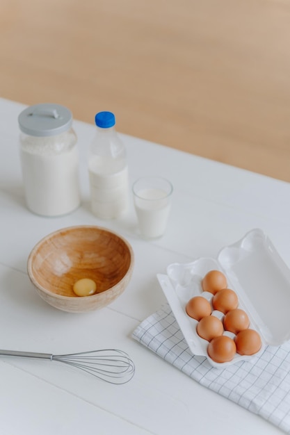 Ingredients for making dough or pasrty Fresh eggs milk and flour with whisk on white wooden kitchen table Products for cooking Vertical shot