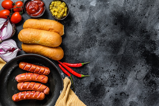 Ingredients for making homemade hot dogs. Sausages in pan, fresh baked buns, mustard, ketchup, cucumbers. Black background. Top view. Copy space