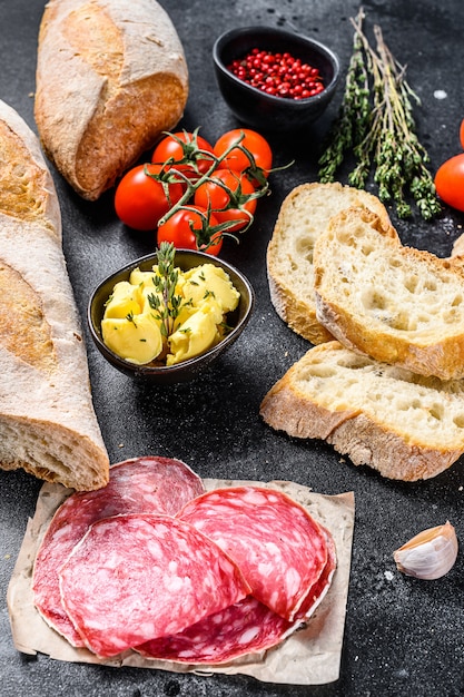 Ingredients for sandwich with butter, smoked meat, baguette, cherry tomatoes, parmesan cheese, garlic and spices. black background. top view.