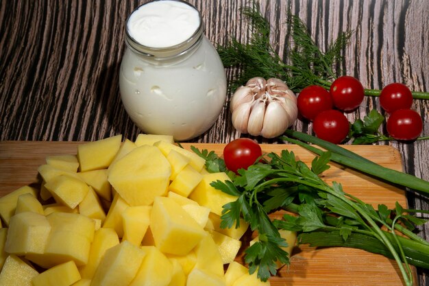 Ingredients for soup or roast sliced raw potatoes on a cutting board next to vegetables and sour cre...