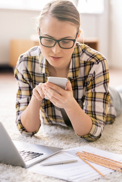 Inhoud peinzende jonge vrouw in glazen liggend op een tapijt en het gebruik van smartphone tijdens het beantwoorden van sms tijdens freelance werk thuis