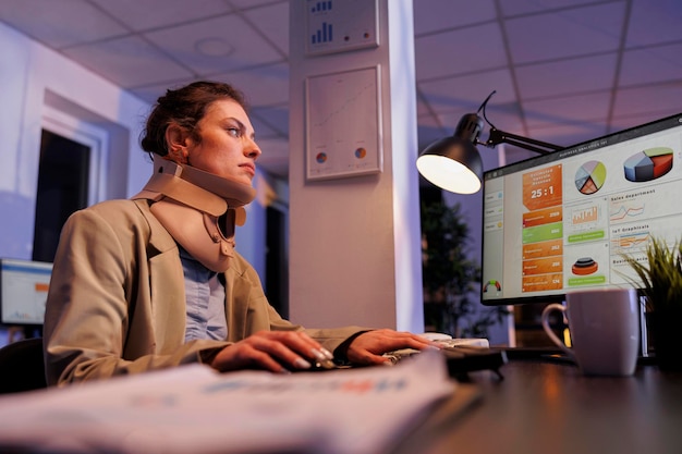 Injrued businesswoman analyzing financial growth report, working late at night at company strategy. Executive manager wearing cervical neck collar in startup office. Corporate concept